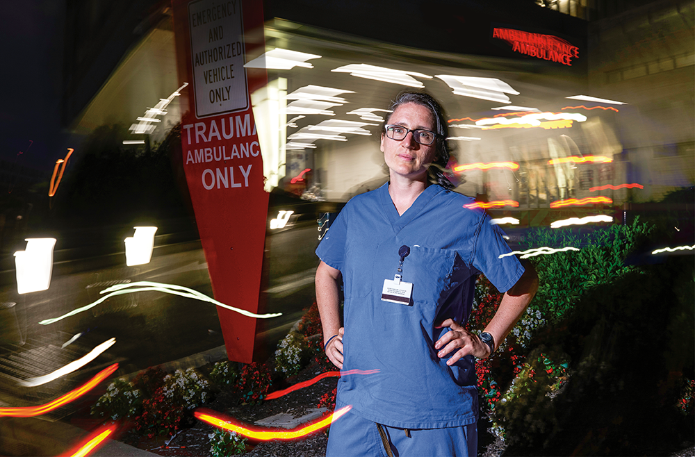 Elinore Kaufman, MD, wearing dark blue scrubs, stands outside the Emergency entrance of Penn Presbyterian Medical Center at night near a sign that says Trauma Ambulance Only.
